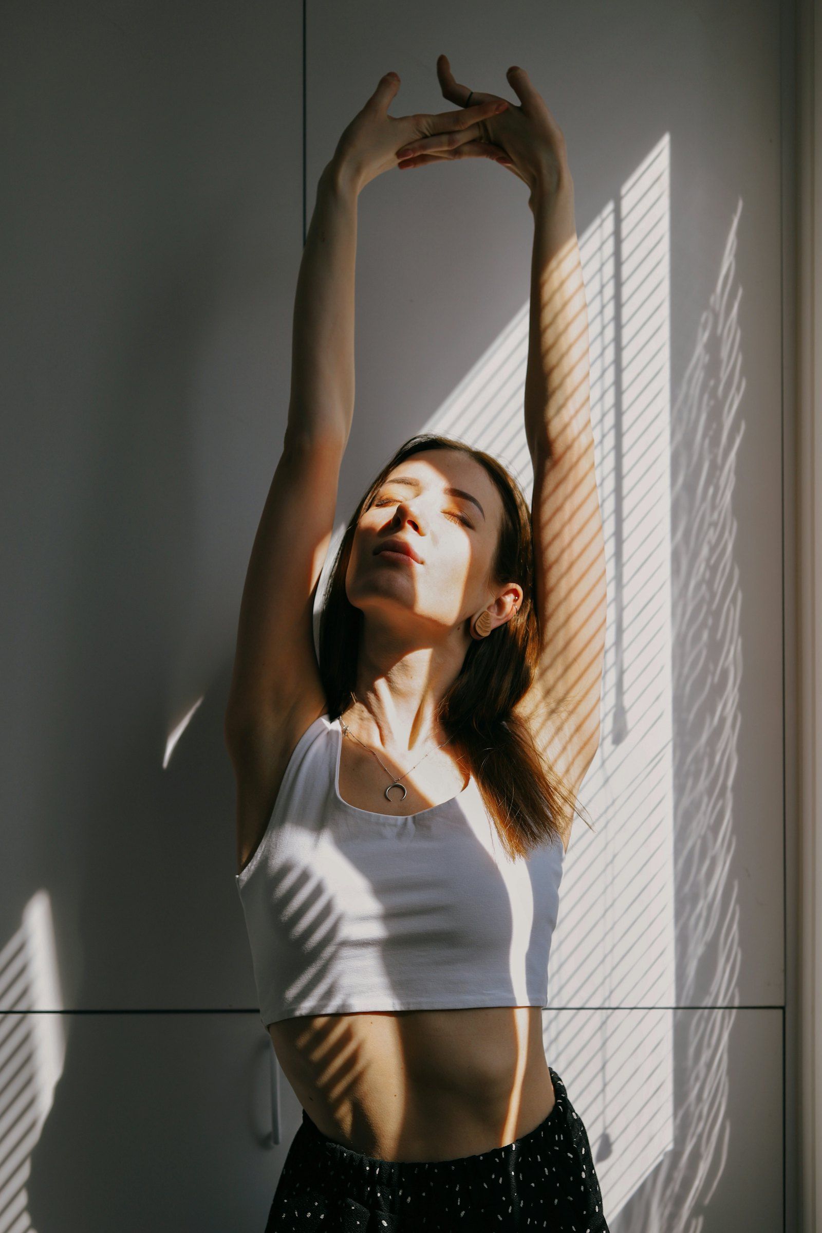 A woman with eyes closed, face turned towards morning sunlight by a window