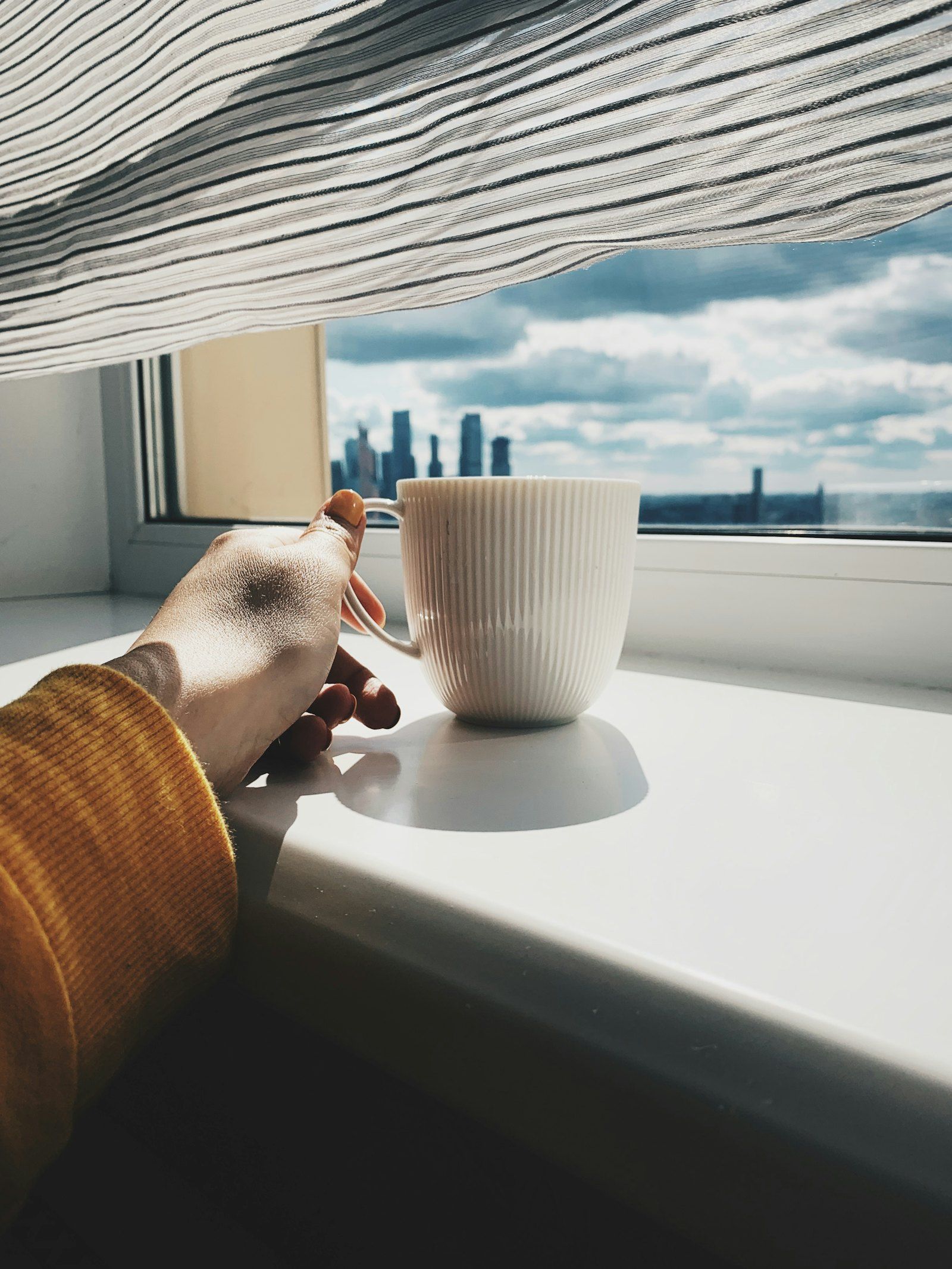 A hand resting beside a ribbed white mug on a sunny windowsill